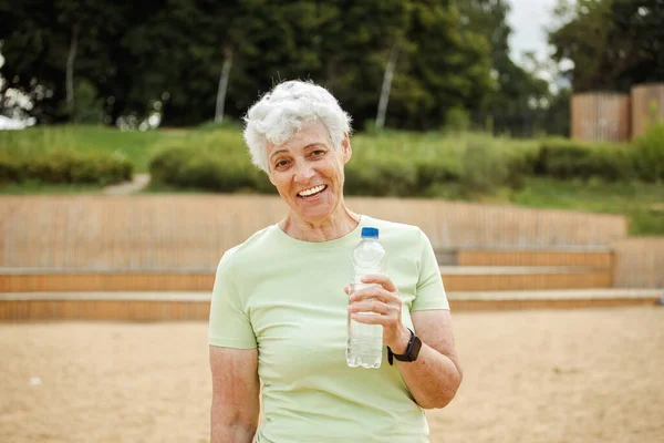 Elderly smiling woman with short grey hair drinking water after exercising, portrait in the park