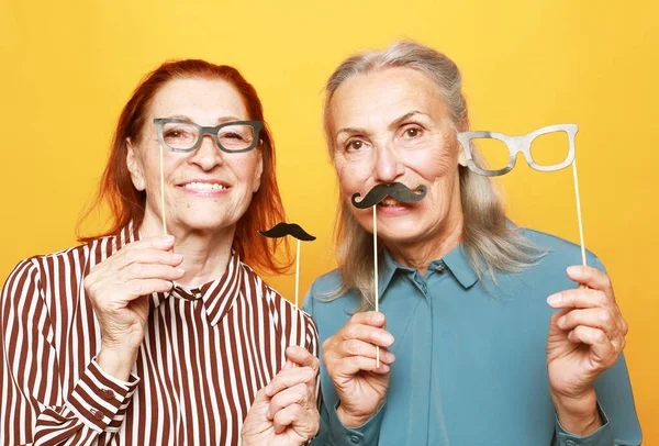 funny smiling elderly female friends with fake mustache and glasses, laughs and prepares for party over yellow background