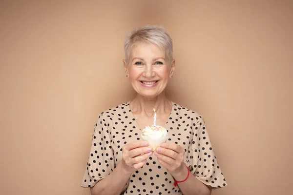 Cheerful elderly woman with short haircut holds a cupcake with a candle in her hands, celebrates a birthday and laughs