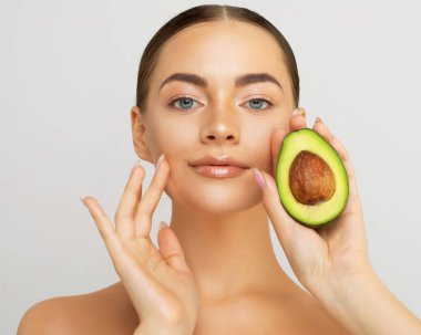 Pretty woman holds half an avocado in front of her face. Photo of attractive woman with perfect makeup on grey background. Beauty and Skin care concept.