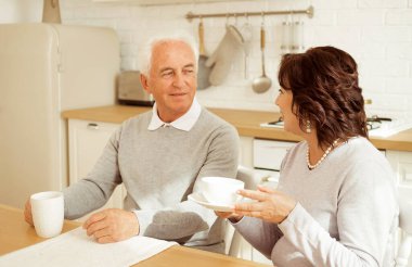 Happy elderly couple drinking coffee in the kitchen while sitting at the table. Family bonds, love and affection concept.