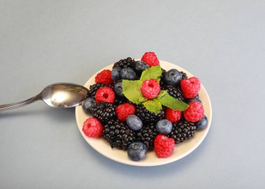 Beyaz tabakta çeşitli meyveler. Yaban mersini, böğürtlen ve ahududu lezzetli diyet meyveleridir. Gri arka planda kaşık ile tabak.