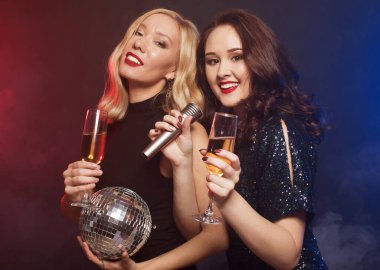İki bayan arkadaş köpüklü şarapla flüt tokuşturuyor ve mikrofon ve disko topları tutuyor. Parti zamanı.