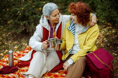 İki yaşlı kadın ormanda ekose bir battaniyeye oturmuş kahve içiyor, konuşuyor ve iyi vakit geçiriyorlar. Yaşam tarzı ve kadın arkadaşlık kavramı.