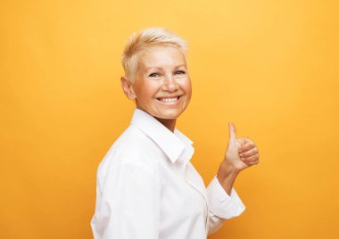 Hayat sadece yaşlandığında başlar. Çekici, mutlu ve tasasız Avrupalı, sarışın, kısa saçlı yaşlı bir kadının portresi gülüyor ve sarı arka planda baş parmağını gösteriyor.