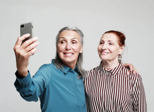 两名老年女性朋友带着一个带着灰色背景手机的自拍 — 图库照片