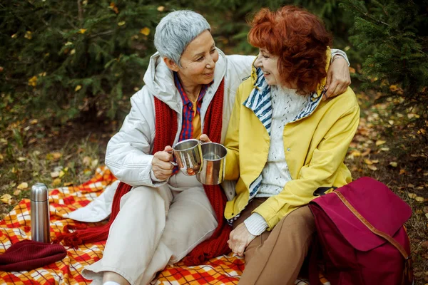 两名老年妇女坐在森林里的一条格子毯子上 一边喝咖啡 一边聊天 玩得很开心 生活方式和女性友谊概念 — 图库照片