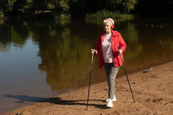Grey-haired woman wearing red jacket walking with tracking sticks on the beach near lake, summer day