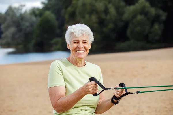 Old woman wear sportswear doing exercises outdoor on the beach in the morning using resistance rubber bands. Healthy lifestyle, active retired life and sporty time.