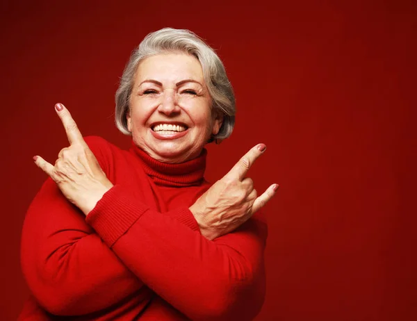 Portrait of funny senior gray-haired woman wearing red sweater and glasses doing a rock and roll symbol over red color background