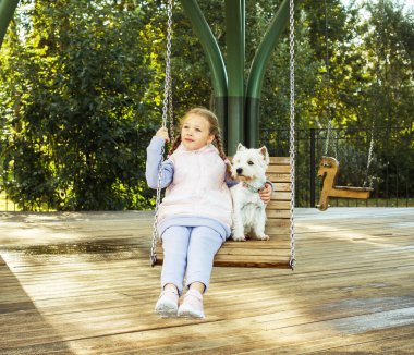 Kız parkta beyaz bir köpekle salıncağa biniyor.