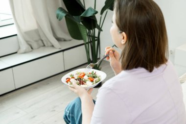 Pencerenin yanında kocaman, mutlu bir kadın ve taze sebze salatası yiyor.