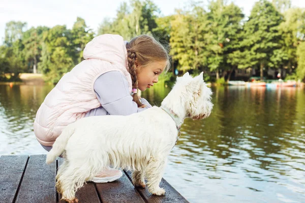 Syvårig Pige Med Lille Hvid Hund Står Bro Ved Søen - Stock-foto