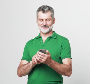 Tehnoloji ve yaşlılar konsepti: Gri arka planda akıllı telefondan mesaj atan yaşlı sakallı adamın portresi, kapatın