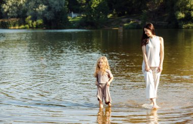 A five-year-old cheerful girl and her mother are walking by the lake, barefoot in the water, having fun on a summer day. Happy time.