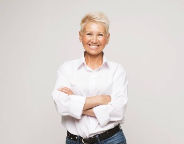 Gri arka planda kolları bağlı yaşlı bir kadın ayakta duruyor. Yaşlı, son sınıf öğrencisi, 60 'lı yaşlarda, beyaz saçlı, kollarını kavuşturup, portreye yakın duran bir kadın..