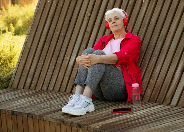 公園内の木製のベンチに座って ヘッドフォンやライフスタイルのコンセプトで音楽を聴く高齢者の女性 — ストック写真