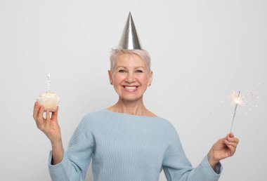 Happy middle aged female wearing conical hat celebrating birthday with cupcake with candle and sparkler over grey background