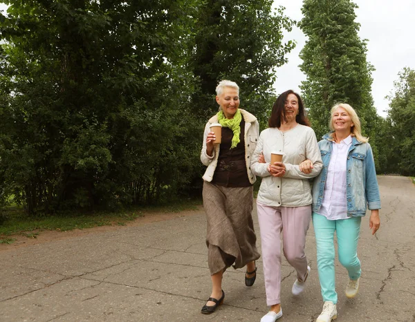 Lifestyle, emotion, old people and gold age concept. Three beautiful elderly women are walking in the park, having fun talking and drinking coffee. Summer day. Happy retirement. Full length portrait.