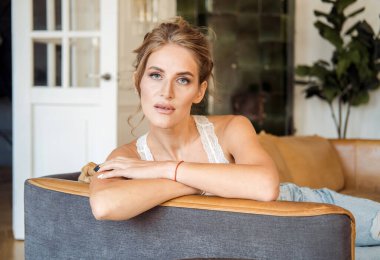 Beyaz dantelli, sarışın, güzel bir kadın oturma odasındaki kanepede oturuyor.