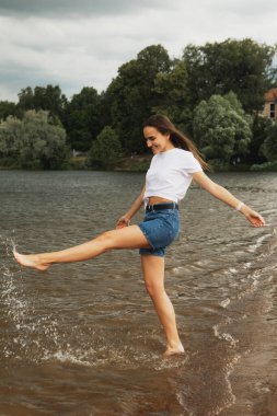 Uzun saçlı neşeli genç kadın göl kenarında yalınayak yürüyor. Kıyıya sıçrayan.