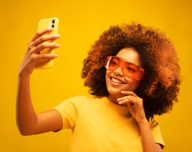 Siyah tenli melez kadın, Afro saç stili, cep telefonu var, video görüşmesi yapıyor, günlük kıyafet ve turuncu güneş gözlüğü takıyor, sarı stüdyo duvarında izole edilmiş..