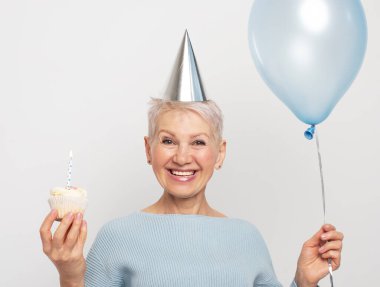 Mutlu yaşlı kadın koni şapkası takıyor doğum gününü mum ve gri arka planda mavi balonla kutluyor.
