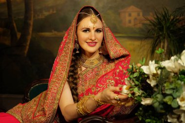 Charming Indian bride dressed in Hindu red traditional wedding clothes embroidered with gold and a veil smiles tender sitting on sofa near flowers.