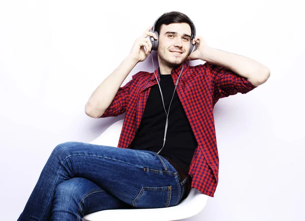 Smiling handsome male model sits in a chair and listens to music in headphones, portrait on a white background. Lifestyle and people concept.
