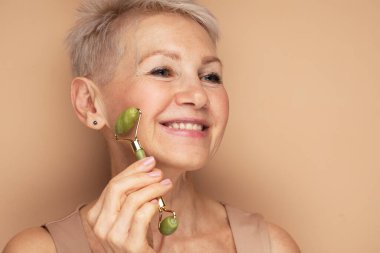 60 'lı yaşlarda güzel bir kadın. Yüzüne masaj yapıyor. Renkli arka planda yeşil kuvars silindiri kullanıyor..