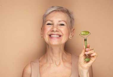 60 'lı yaşlarda güzel bir kadın. Yüzüne masaj yapıyor. Renkli arka planda yeşil kuvars silindiri kullanıyor..