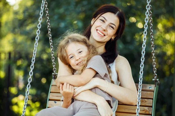 Mom and daughter swing on a swing. Caucasian woman and little girl have fun on the playground. Lifestyle and people concept.