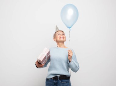 İnsanlar, yaşlanma ve olgunluk kavramı. Kısa saçlı, parti şapkası takan, elinde mavi balon ve beyaz gri arka planda hediye kutusu olan yaşlı bir kadın. Kameraya bakıyorum..