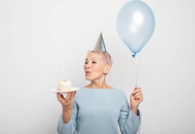 İnsanlar, parti, yaşlanma ve olgunluk kavramı. Doğum gününü kutlayan koni şapkalı, elinde mum ve mavi balonla izole bir şekilde poz veren orta yaşlı, güzel bir kadın. Mutlu zamanlar..