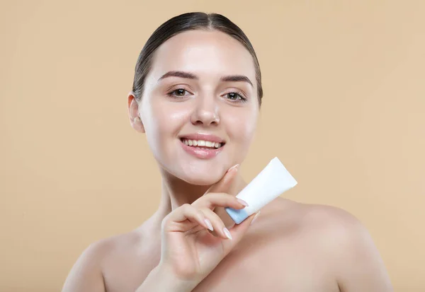 Beauty Youth Skin Care Concept - Beautiful Caucasian Woman Face Portrait holding and presenting cream tube product. Beautiful Spa model female with Perfect Fresh Clean Skin over beige background.
