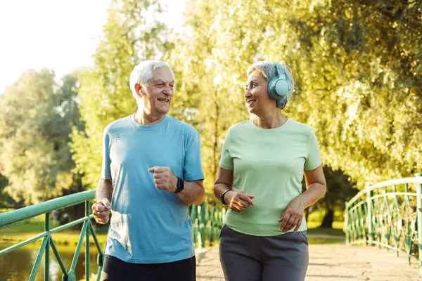 Senior mature couple running together in the park looking at each other while jogging slimming exercises. Training workout
