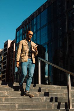 Genç Afro adam güneş gözlüğü takıyor. Modern iş merkezinin yakınına mesaj göndermek için akıllı telefon kullanıyor. Yaşam tarzı ve insan kavramı.