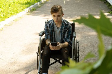 Tekerlekli sandalyedeki genç bir adam elinde akıllı telefon tutuyor ve haberleri okuyor. Parkta yaz günü