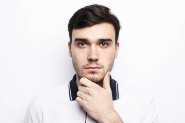 Genç esmer adam beyaz tişört giyer ve gri arka plandaki kameraya bakar.