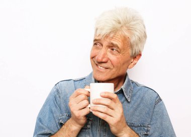 Yaşlı, neşeli, yaşlı bir adam elinde bir kupa kahveyle beyaz arka planda gülümsüyor.