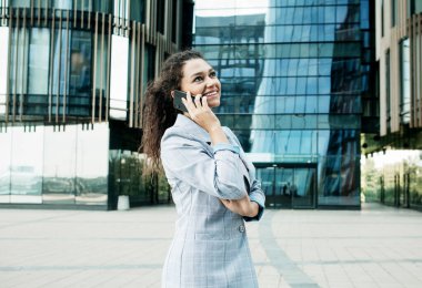 İş merkezinin arka planında iş tarzı giyinmiş genç bir Afrikalı kadın elinde bir cep telefonu tutuyor.