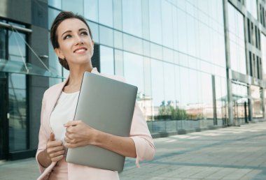 İş, iletişim, teknoloji ve insan konsepti - arka planda ofis binaları ile sokakta dizüstü bilgisayarı olan genç bir iş kadını