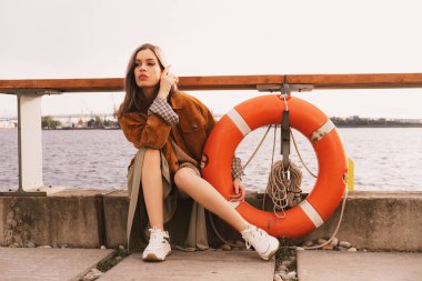 Şık ve şık bir kadın model nehir kenarındaki setin yanında bir can simidinin yanında oturuyor.