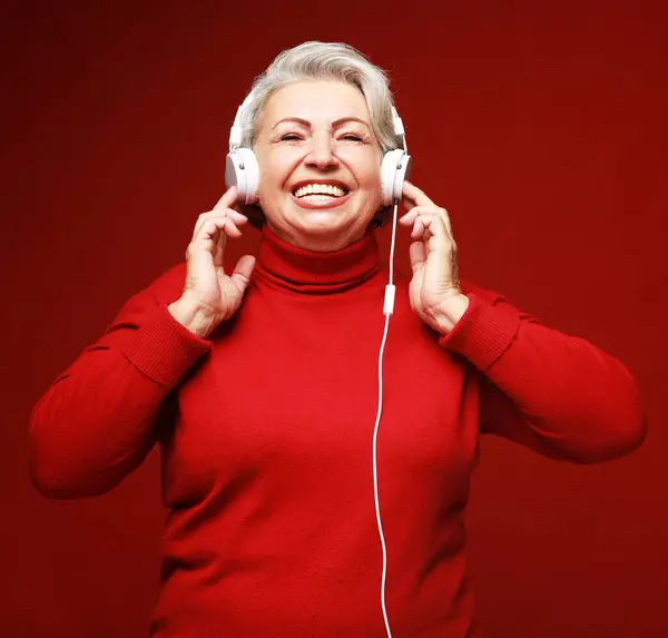 Lifestyle, party, emotion and old people concept: elderly woman wearing red sweater listening to music with headphones