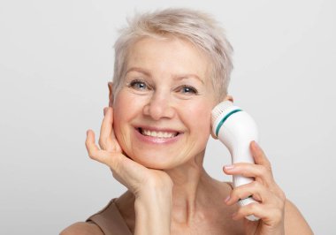 Gri arka planda mükemmel bir cilt için yüz masajı yapan kısa saçlı tatlı ve güzel yaşlı bir kadının portresi.