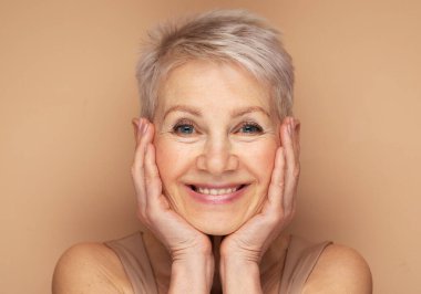 Elleri yüzlerinde gülümseyen olgun bir kadının güzel portresi. Bej arkaplan üzerindeki portreyi kapat.