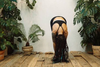 Saçları örgülü, güzel, hoş ve sportif bir kadın, parlak odada egzersiz yapıyor. Yerde oturup yoga yapan esmer kıza odaklanmış. Ev havası, çiçekler, yaşam tarzı.