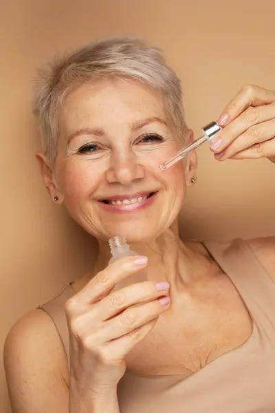 Happy senior lady applies cosmetic oil serum on face takes care of skin and smiles broadly enjoys beauty treatments. Portrait over beige background.