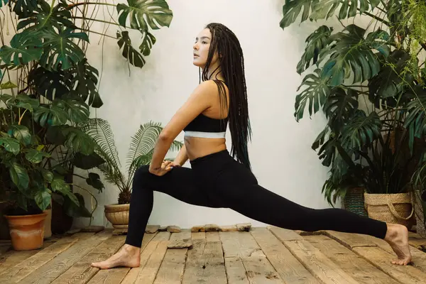 The girl with braids does yoga against the background of green plants in his studio