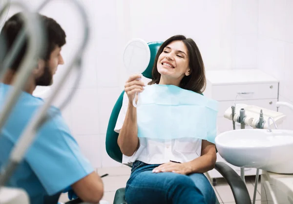 Young female patient holding mirror and looking at his beautiful smile sitting at the dental office. Happy smile.